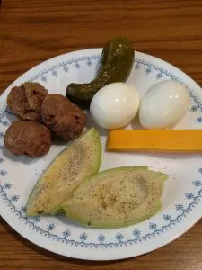 2 avocado quarters, 3 meatballs, 2 hard boiled eggs, cheese stick and pickle on plate