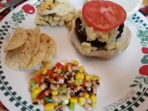 hamburger with onion relish, tomato next to corn and red pepper salsa and chips and potato salad