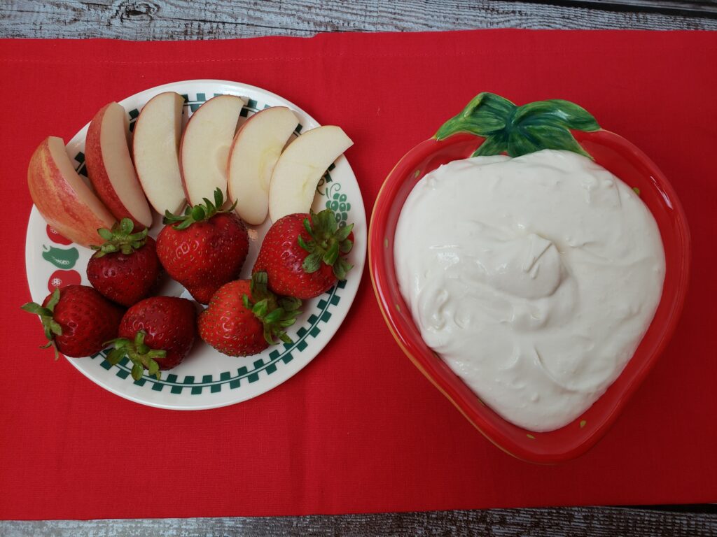 fruit dip in serving bowl next to strawberries and apple slices and on red cloth.