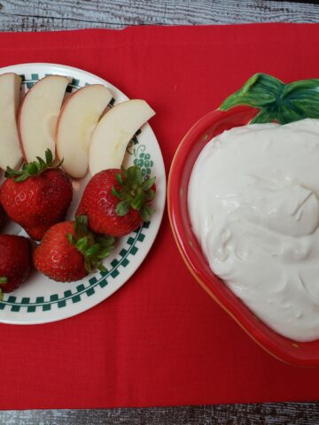fruit dip in serving bowl next to strawberries and apple slices and on red cloth.