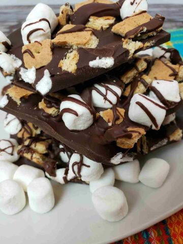 s'mores bark broken and stacked on a white plate with mini marshmallows in front of it.