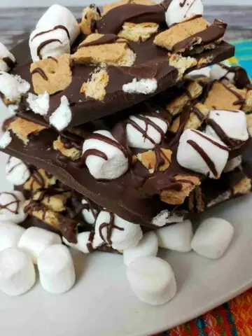 s'mores bark broken and stacked on a white plate with mini marshmallows in front of it.