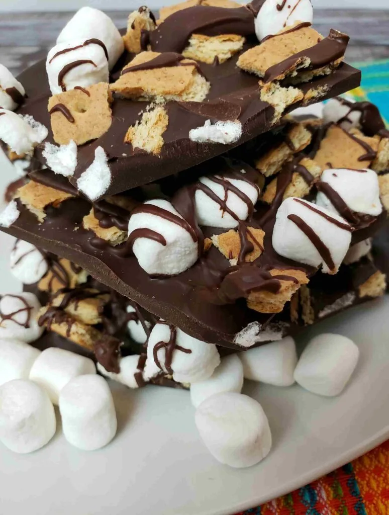 s'mores bark broken and stacked on a white plate with mini marshmallows in front of it.