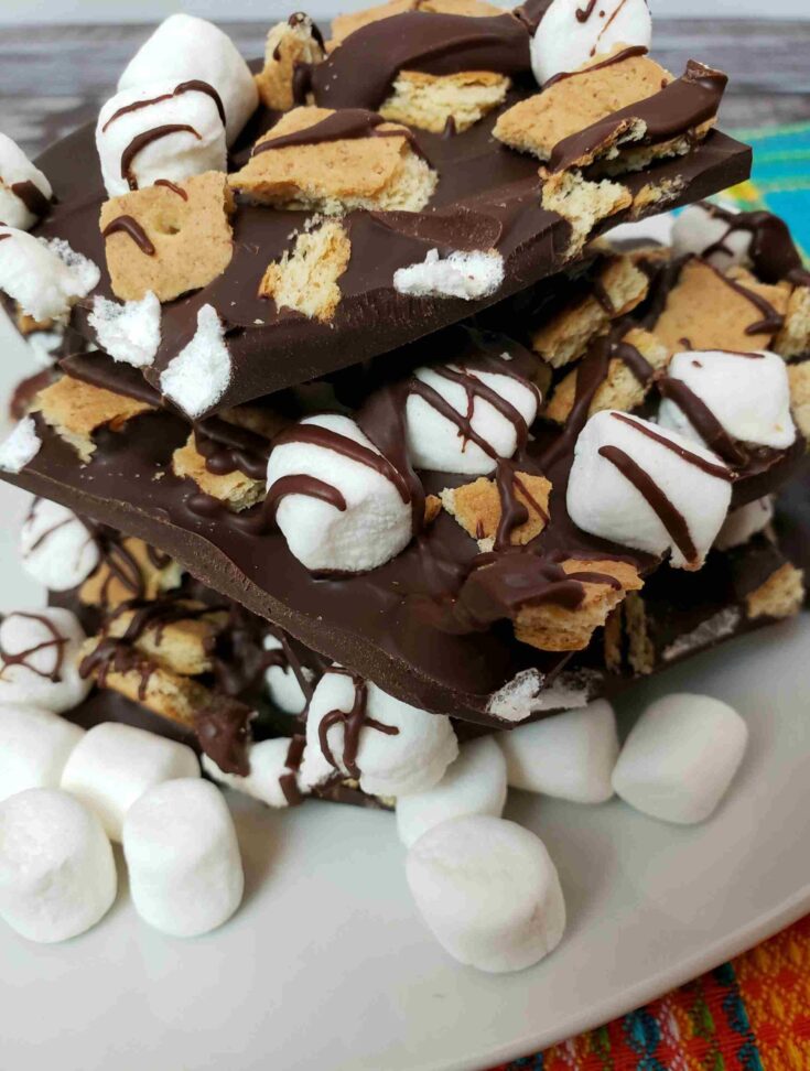 s'mores bark broken and stacked on a white plate with mini marshmallows in front of it.