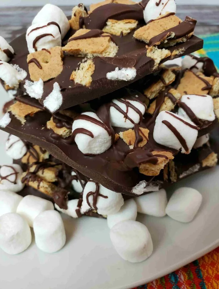 s'mores bark broken and stacked on a white plate with mini marshmallows in front of it.